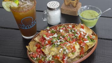 Une assiette de nachos garnie de tomates coupées en dés, d’oignons et de fromage, un verre de thé glacé avec une tranche de citron vert, et un petit bol de sauce verte sur une table en bois.