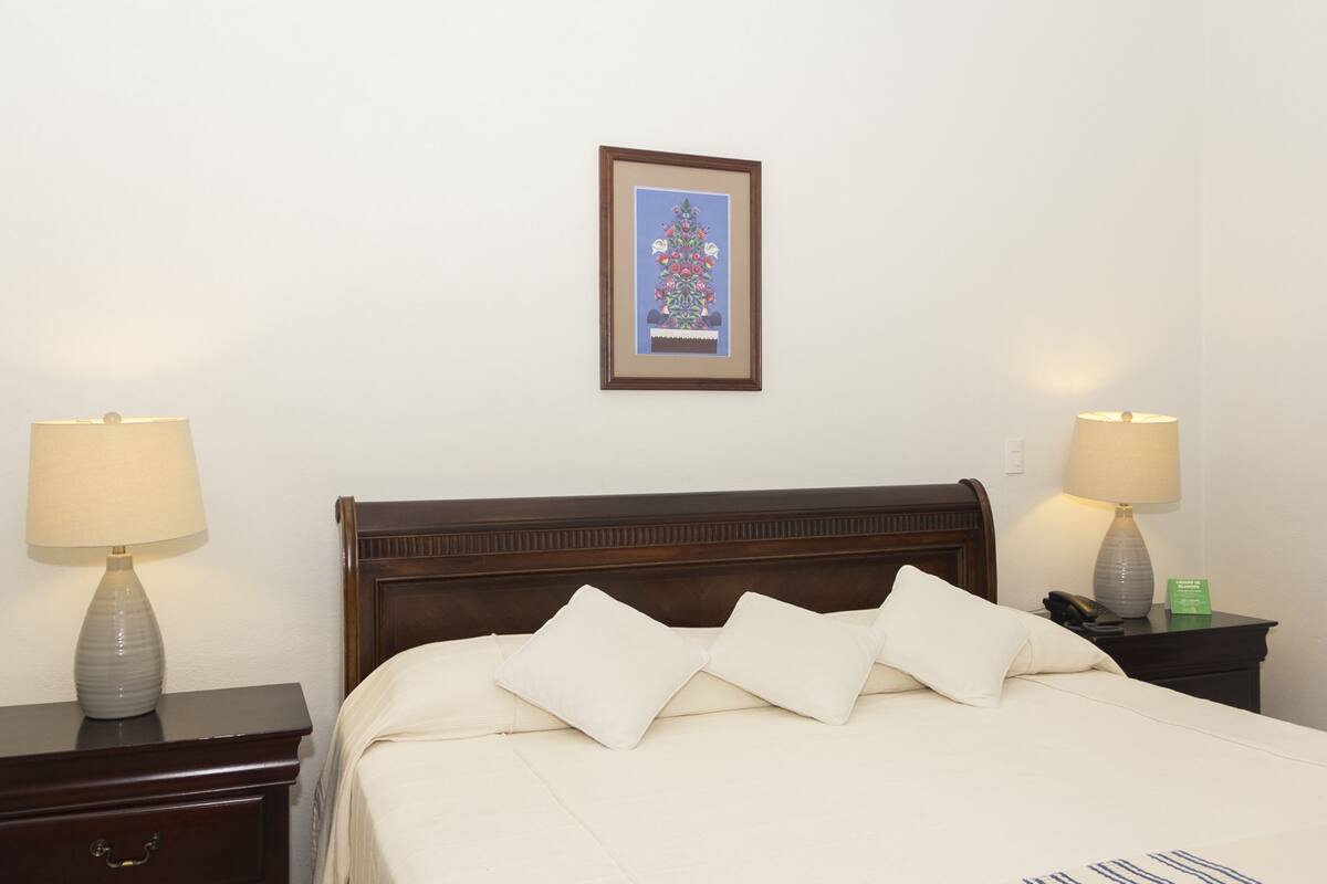 Quarto de hotel com cabeceira de madeira, duas mesas de cabeceira com luminárias, um quadro na parede e cama com roupa de cama e almofadas brancas.