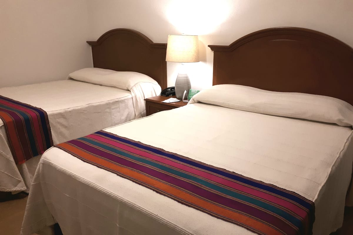 Duas camas de solteiro com roupa de cama branca e colchas listradas coloridas, uma mesa de cabeceira com abajur e um telefone fixado na parede.