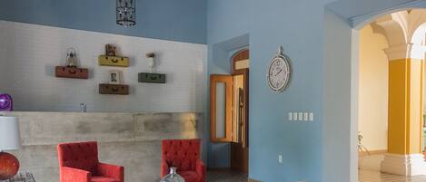 Una sala de estar con dos sillones rojos, una mesa de centro de madera y un sofá color beige. Se observan estantes montados en la pared con objetos decorativos y un reloj de pared.