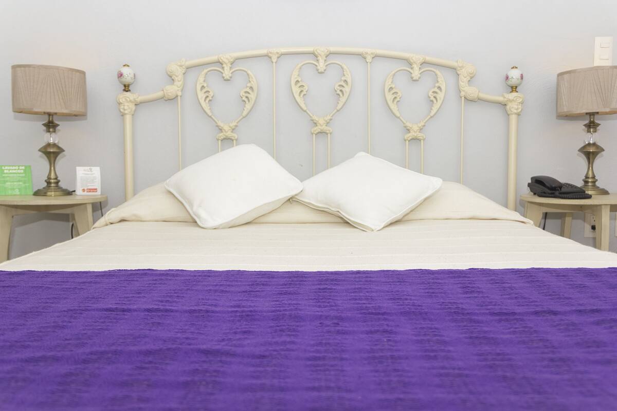 Uma cama com cabeceira branca e duas almofadas, um cobertor roxo e duas mesas de cabeceira com abajures e um telefone.