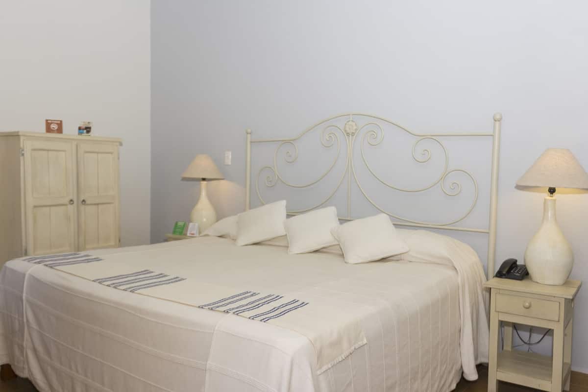 Cama bem arrumada com colcha e travesseiros brancos, cabeceira de metal branca, mesa de cabeceira com abajur e pequeno armário com alguns objetos em cima.