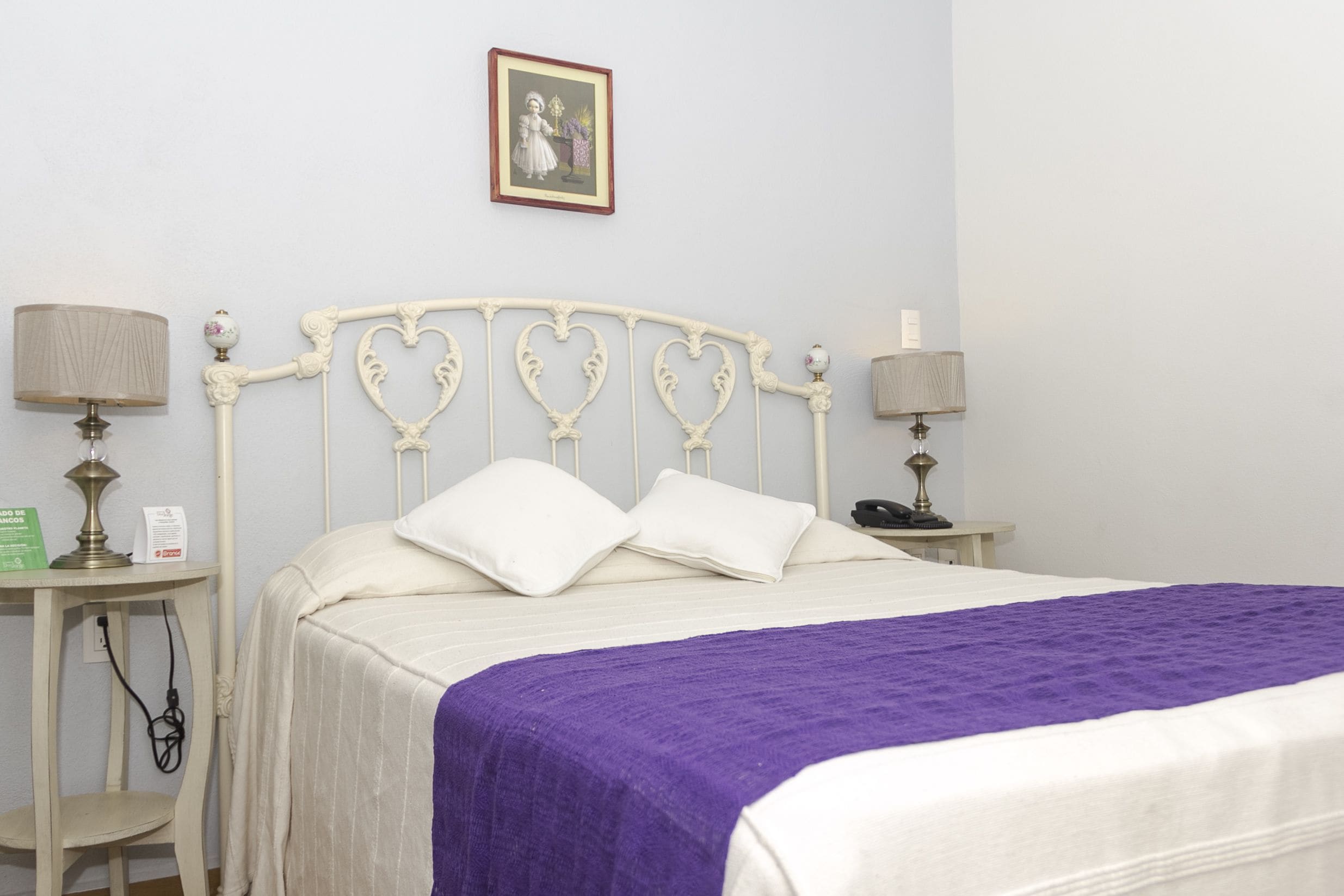 A neatly made bed with a purple blanket, white pillows, and a heart-shaped headboard. There are two bedside tables with lamps and a small shelf with a lamp and a box.