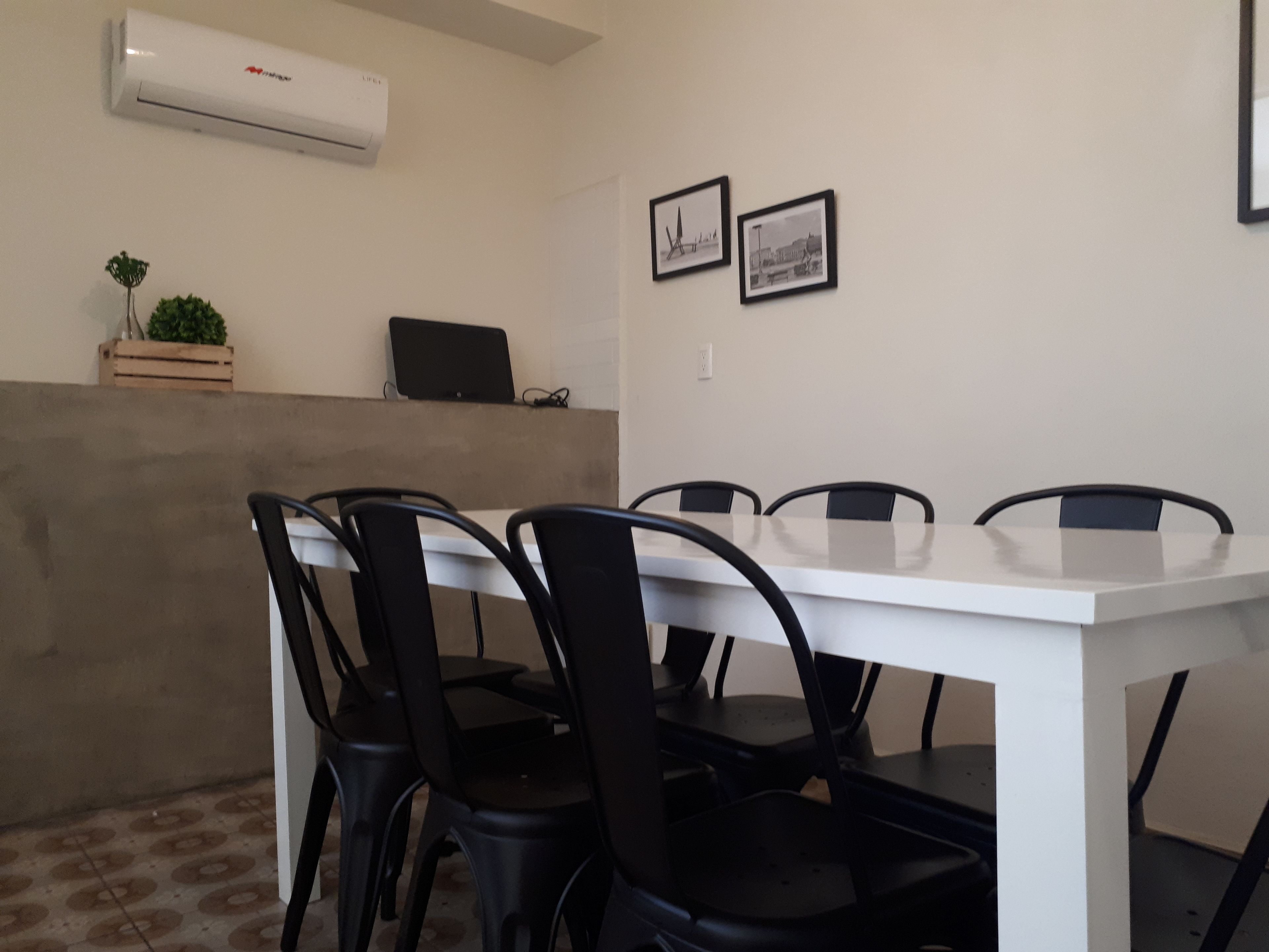 Une salle de conférence avec une table blanche, des chaises noires, un climatiseur, un ordinateur portable et deux tableaux encadrés au mur.