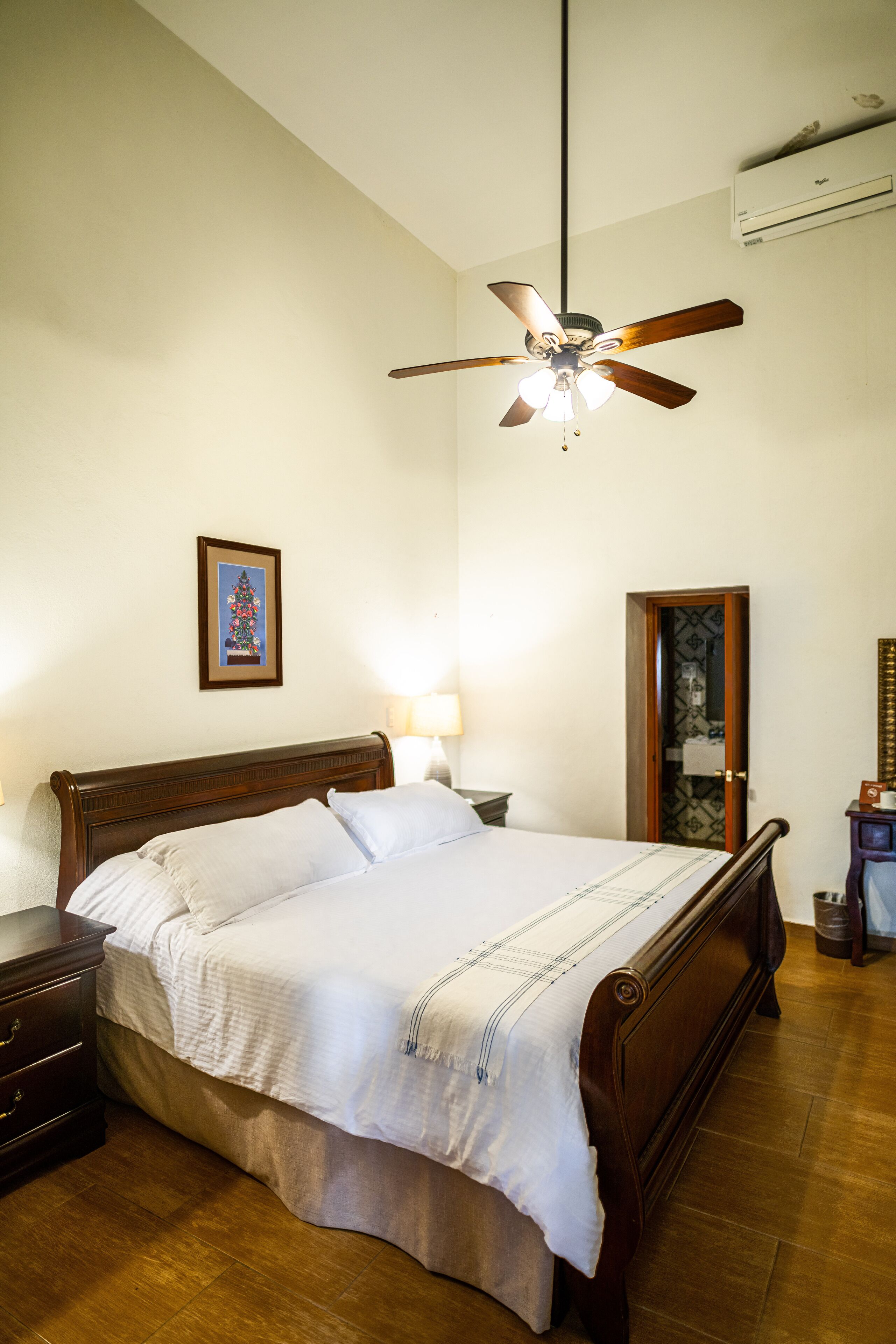 A neatly made bed with a wooden headboard, a ceiling fan, a framed picture on the wall, and a nightstand with a lamp.