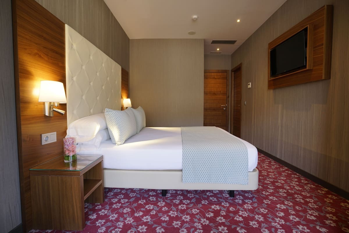 A hotel room with a large bed, a flat-screen TV mounted on the wall, a bedside table with a lamp, and a floral-patterned carpet.