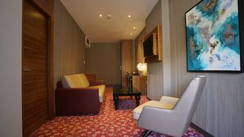 Quarto de hotel com carpete estampado de flores vermelhas, um sofá, uma cadeira, uma TV fixada na parede e um quadro.