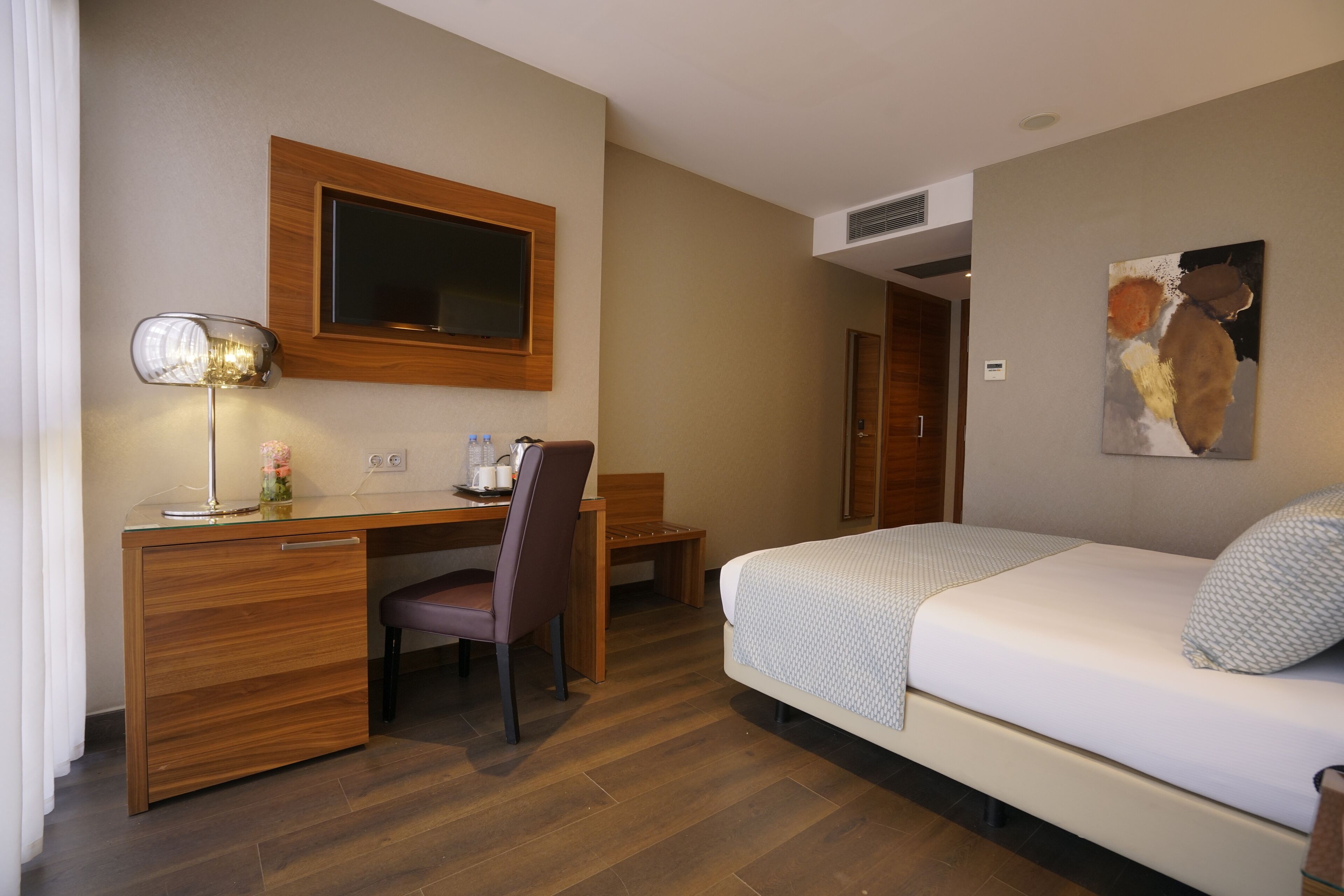 Una habitación de hotel con un escritorio de madera, una silla, una cama con una colcha estampada, un televisor montado en la pared y un cuadro.