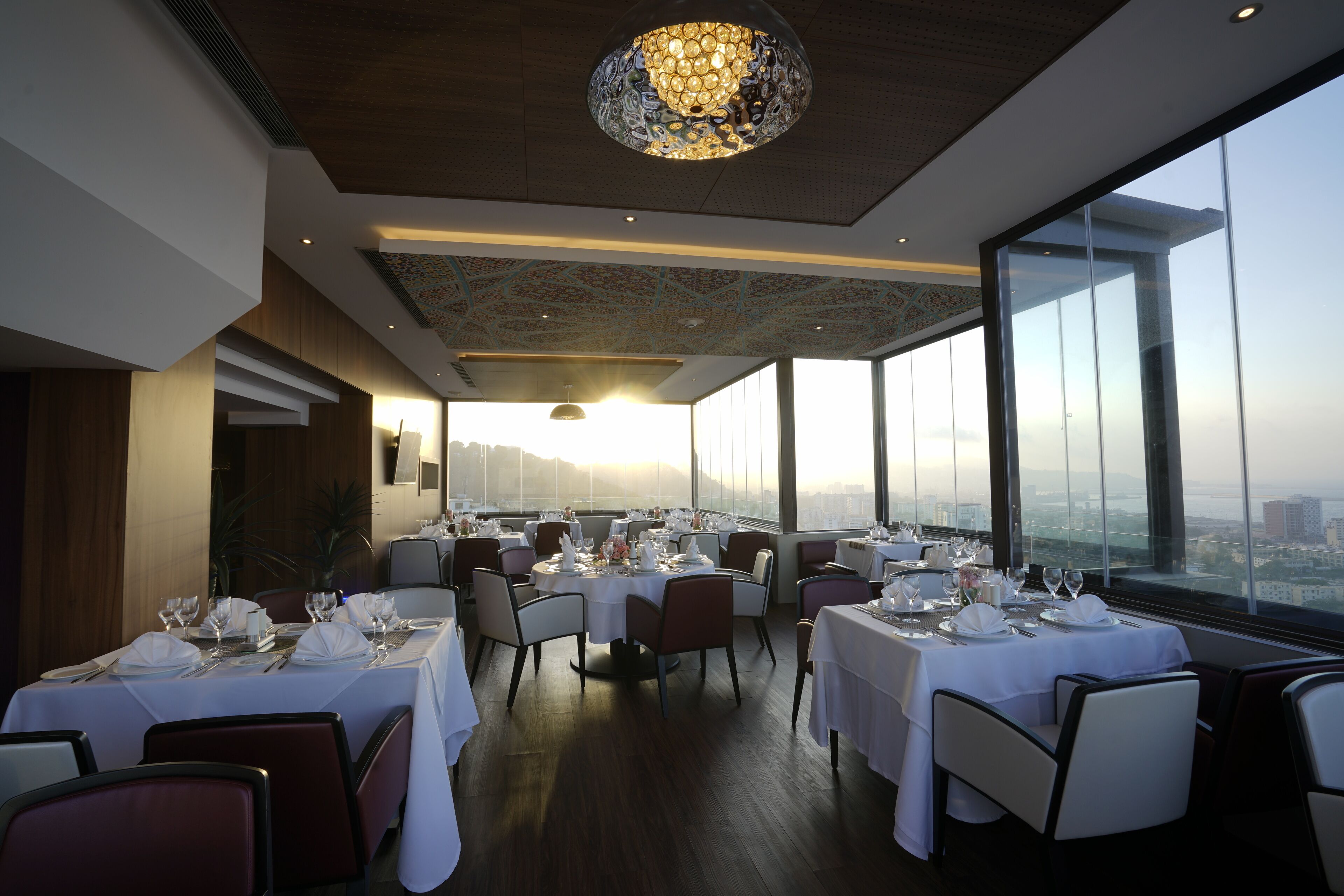 Un restaurante con mesas redondas dispuestas para comer, amplios ventanales con vista a la ciudad y una lámpara decorativa.