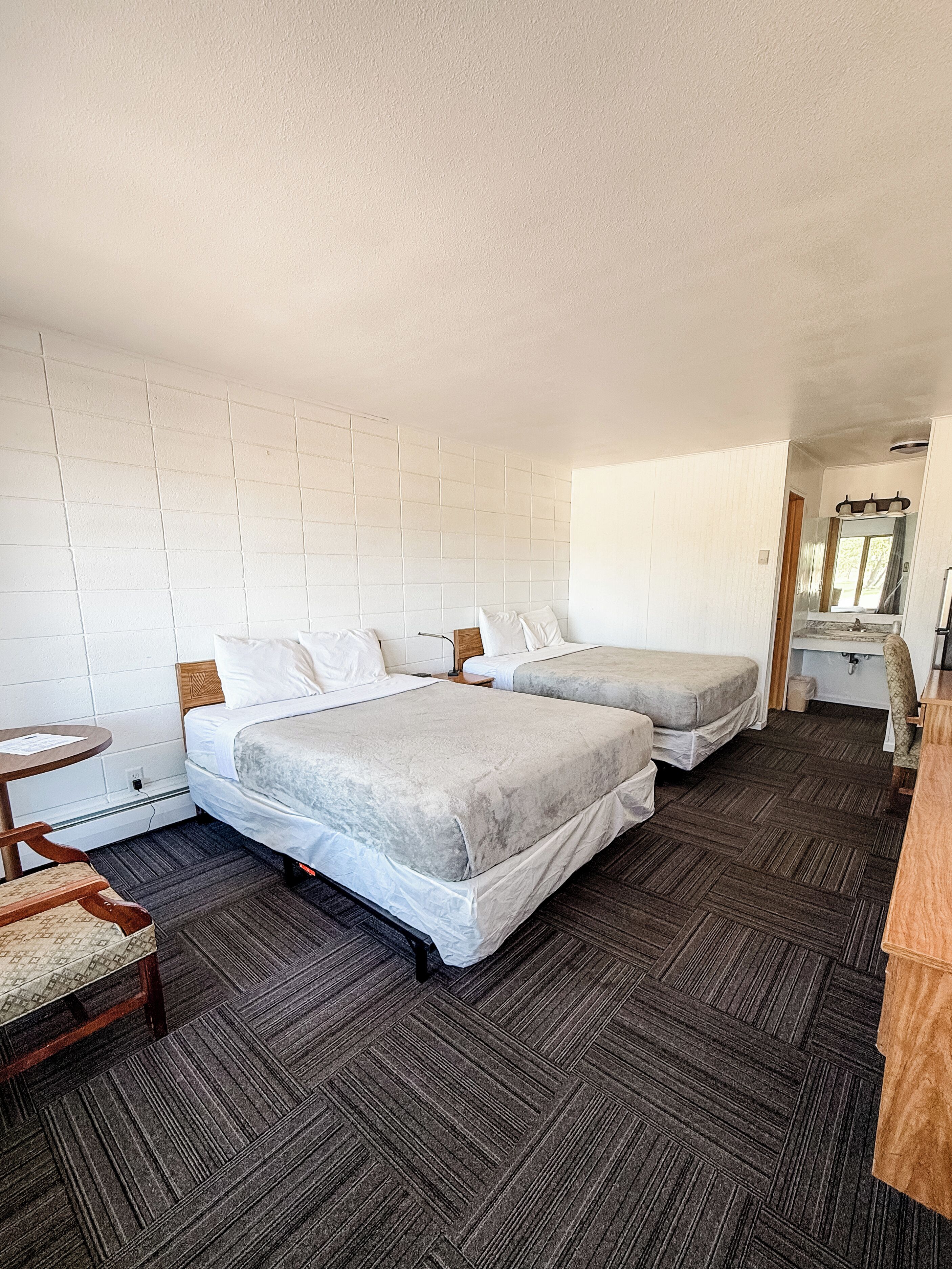 A hotel room with two beds, a small table, a chair, and a bathroom with a sink and mirror.