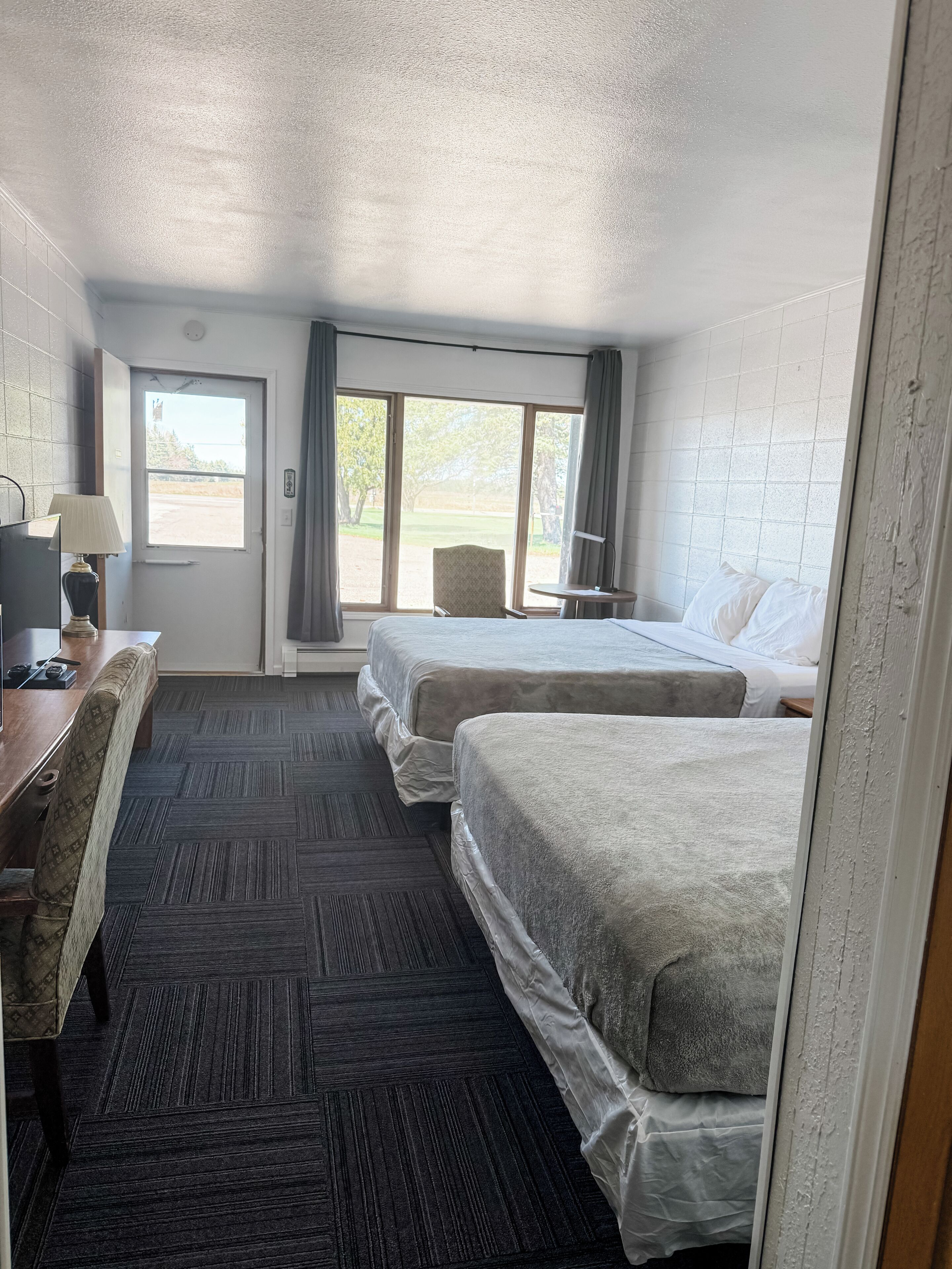 A hotel room with two beds, a desk with a lamp, a chair, and a window with a view of trees and a building.