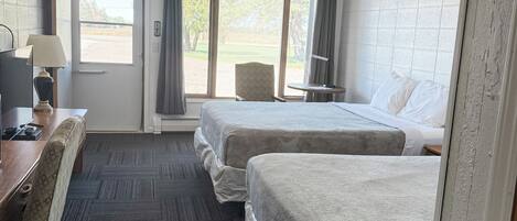 Habitación de hotel con dos camas, un escritorio con lámpara, una silla y una ventana con vistas a árboles y un edificio.