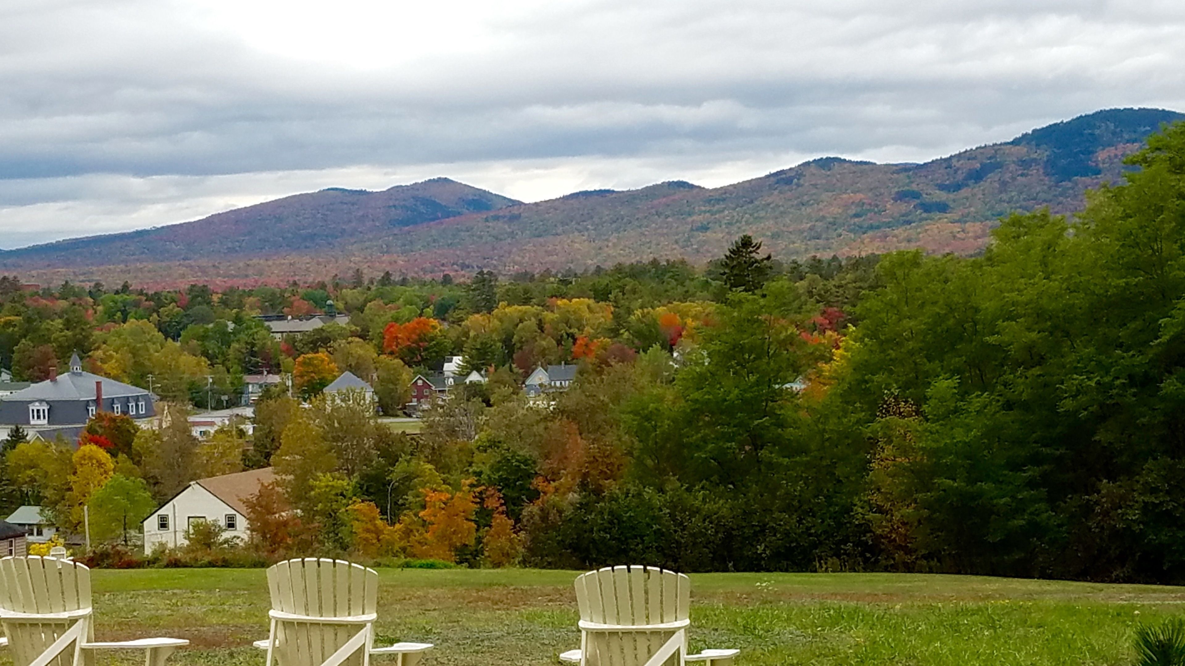A view of a mountainous landscape with autumn foliage, a grassy area with three white Adirondack chairs, and several houses in the foreground.