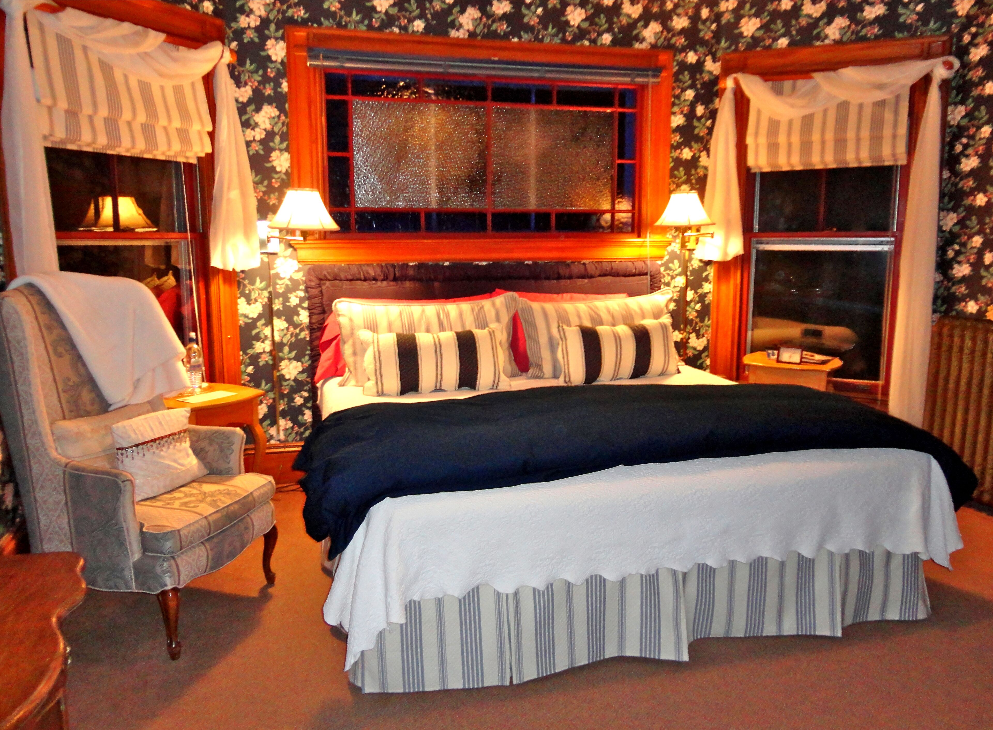 Un dormitorio con una cama grande, dos lámparas de noche, una silla y papel tapiz con motivos florales.
