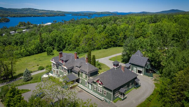 A large, green, two-story house with a spacious front yard and a view of a lake.