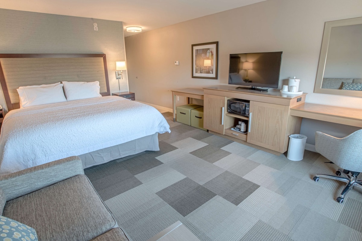 A hotel room with a large bed, a desk with a computer, a TV, and a small kitchenette.