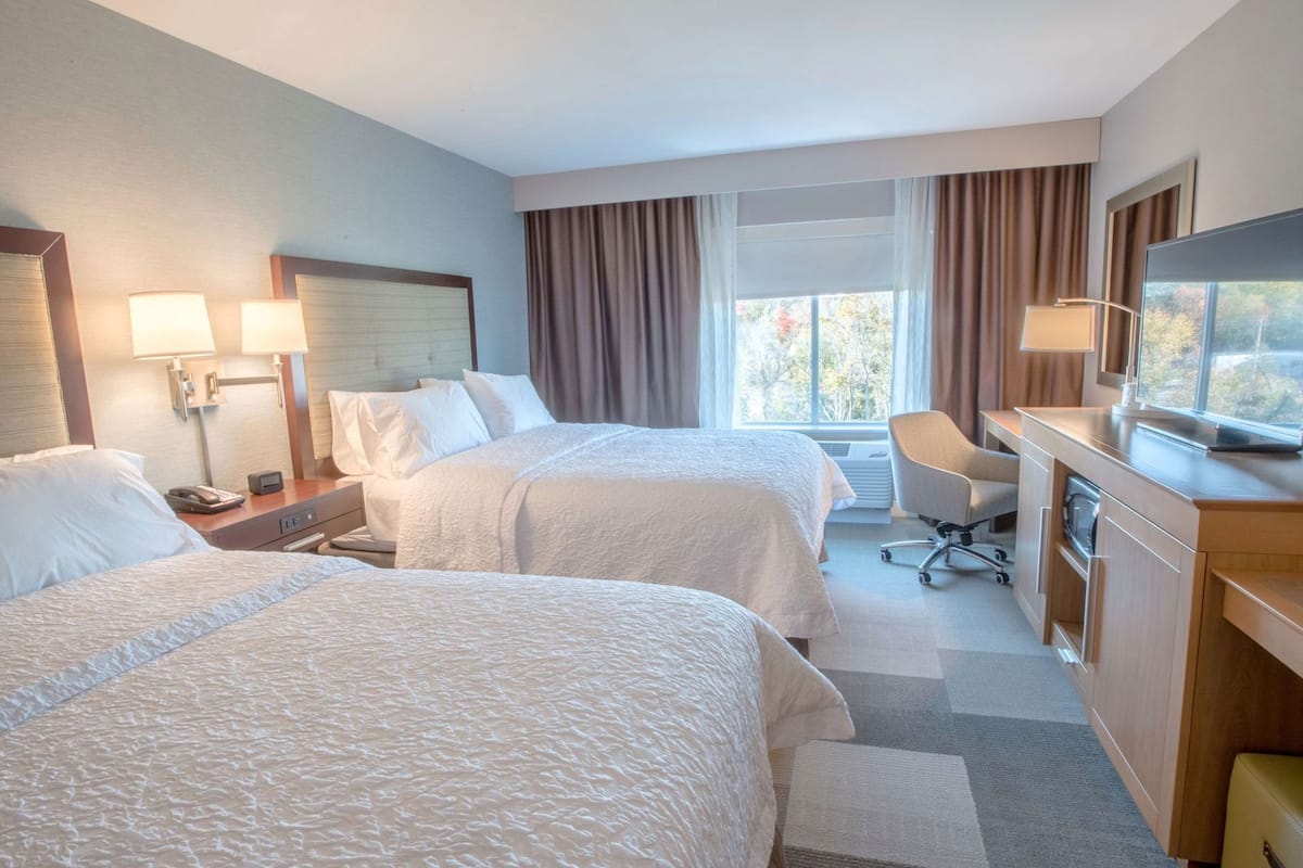 A hotel room with two beds, a desk with a television, a chair, and a window with curtains.