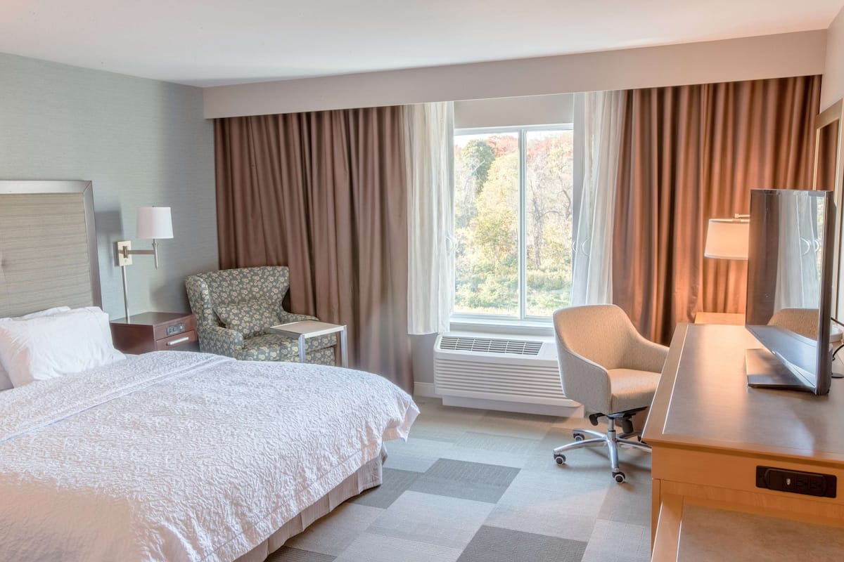A hotel room with a large bed, two armchairs, a desk with a computer, a window with curtains, and a wall-mounted TV.
