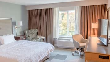 Ein Hotelzimmer mit einem großen Bett, zwei Sesseln, einem Schreibtisch mit Computer, einem Fenster mit Vorhängen und einem an der Wand montierten Fernseher.