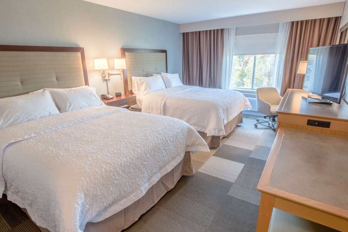 A hotel room with two beds, a desk, a chair, a TV, and a window with curtains.