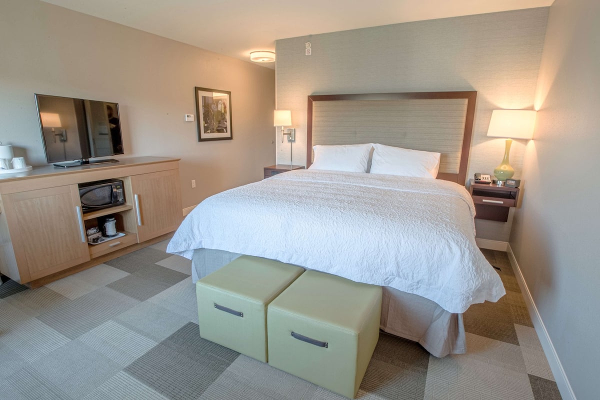 A hotel room with a large bed, a TV, a desk with a lamp, and a small kitchenette.
