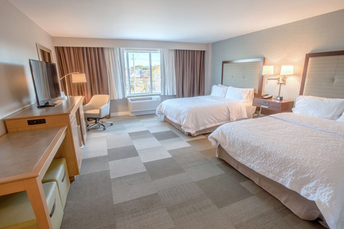 A hotel room with two beds, a desk with a chair, a television, and a window with curtains.