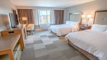 Quarto de hotel com duas camas, uma secretária com cadeira, televisão e janela com cortinas.