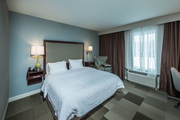 Una habitación de hotel con una cama grande, una mesita de noche con lámpara, una silla, una ventana con cortinas y un equipo de aire acondicionado.