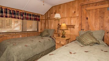 Ein Zimmer mit zwei Betten, Holzwänden, einem Fenster mit Jalousien und einem Nachttisch mit Lampe.