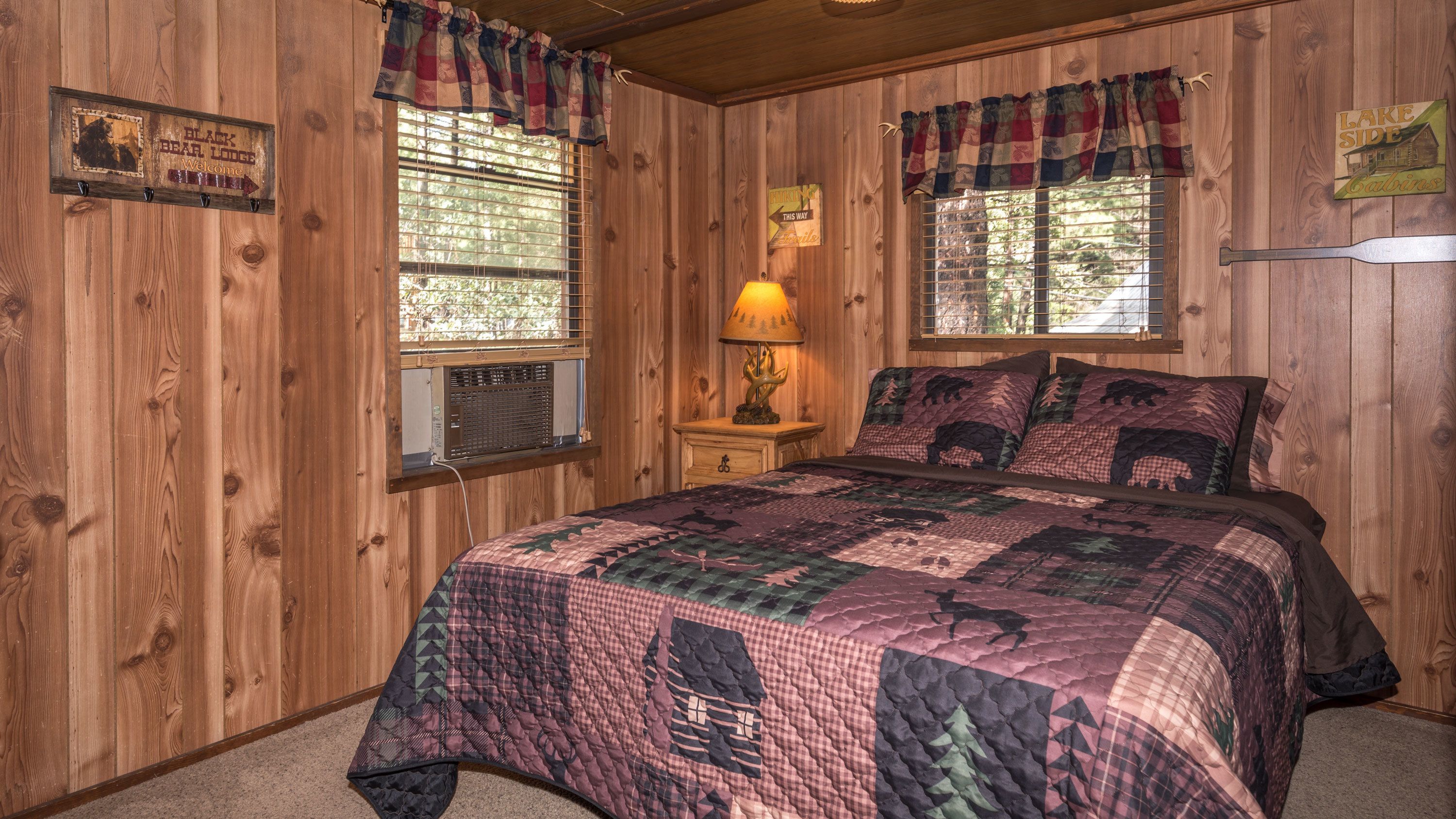 A wooden cabin room with a bed, a nightstand, a lamp, and a sign indicating 'Black Bear Lodge Welcome'.