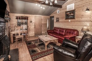 A cozy cabin interior with a wooden floor, a red leather sofa, a black leather armchair, a wooden coffee table, and a fireplace. - Canyon Cabins (Ruidoso)