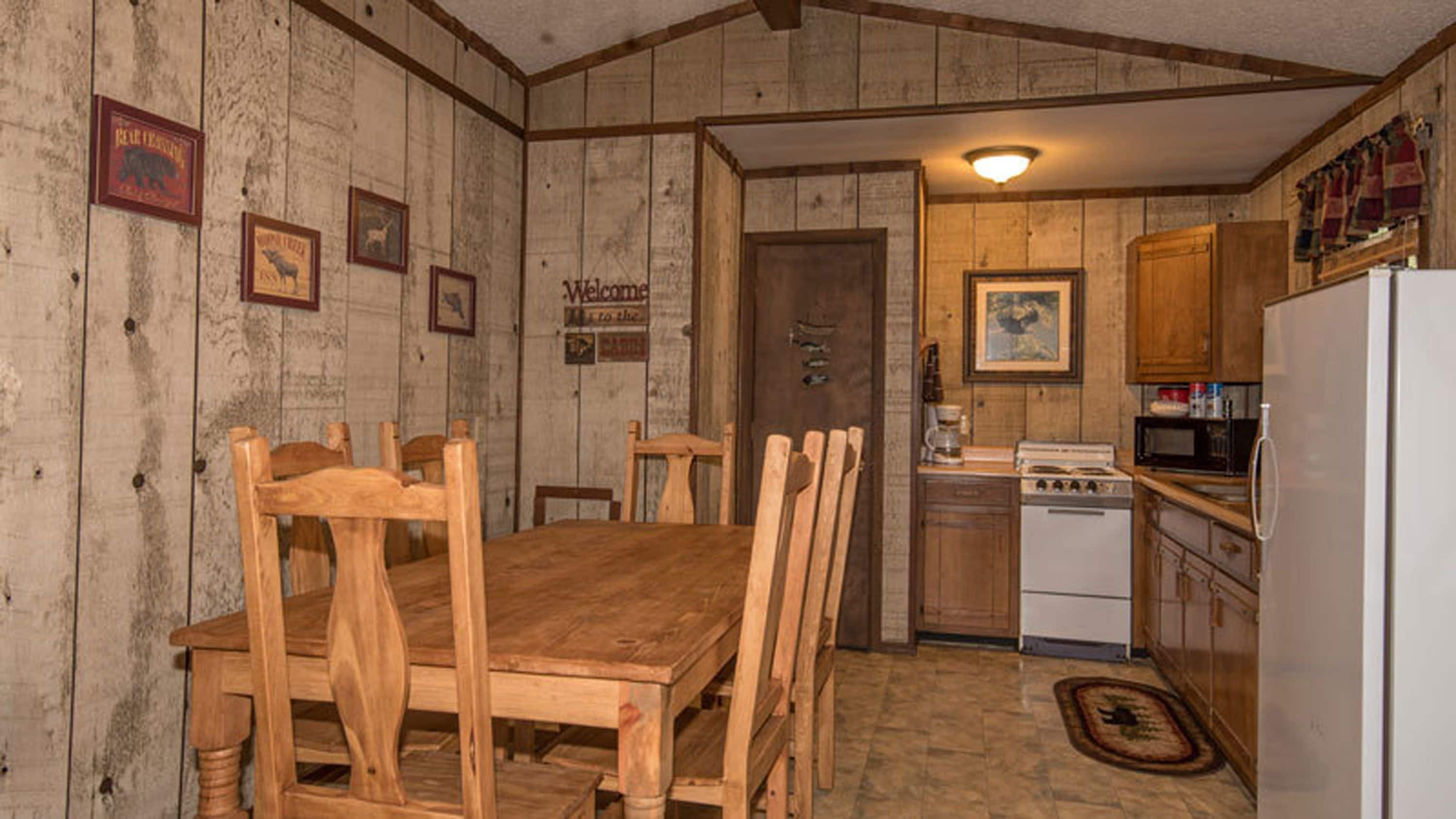 A rustic kitchen with wooden walls, a dining table with chairs, a stove, and a refrigerator.