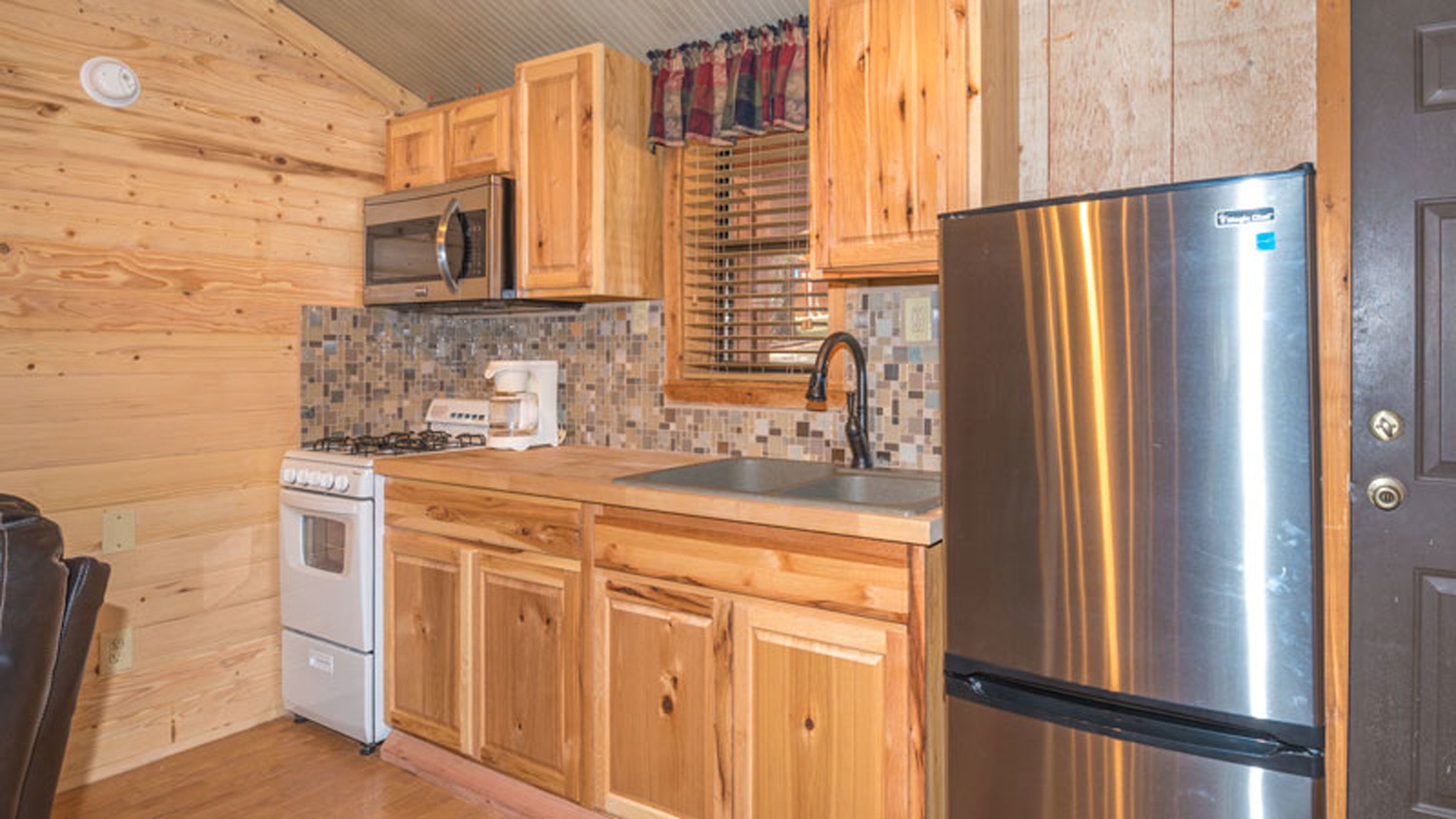 A compact kitchen with wooden cabinets, a stainless steel refrigerator, a microwave, a stove, and a coffee maker.