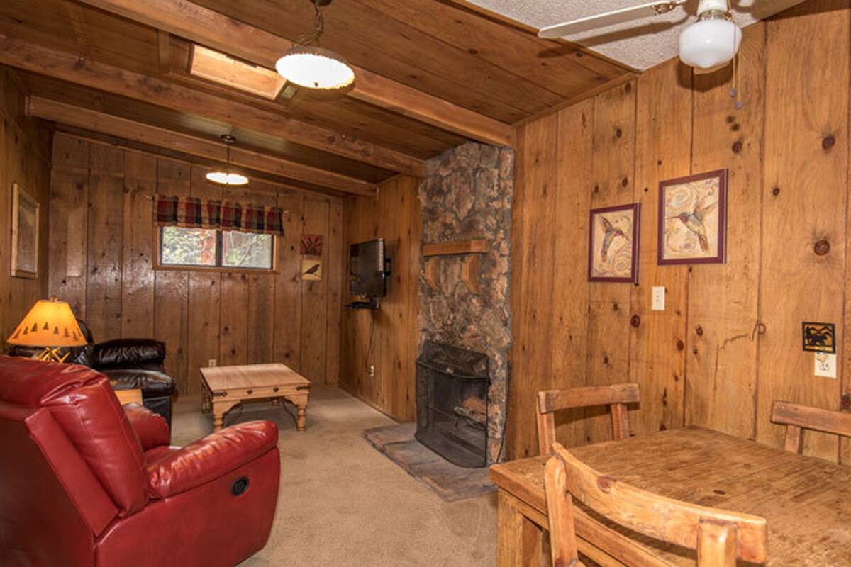 A cozy cabin interior with a stone fireplace, wooden walls, a leather chair, a wooden table, and a television mounted on the wall.