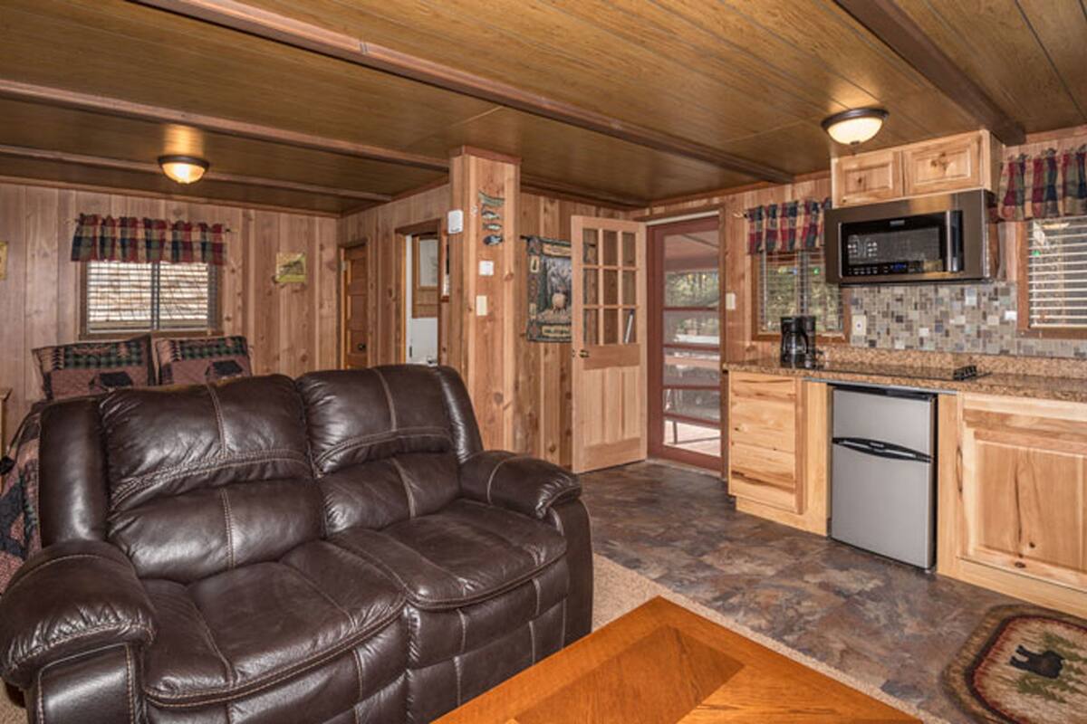 A cozy cabin interior with a leather couch, wooden cabinets, a kitchen area, and a fireplace.