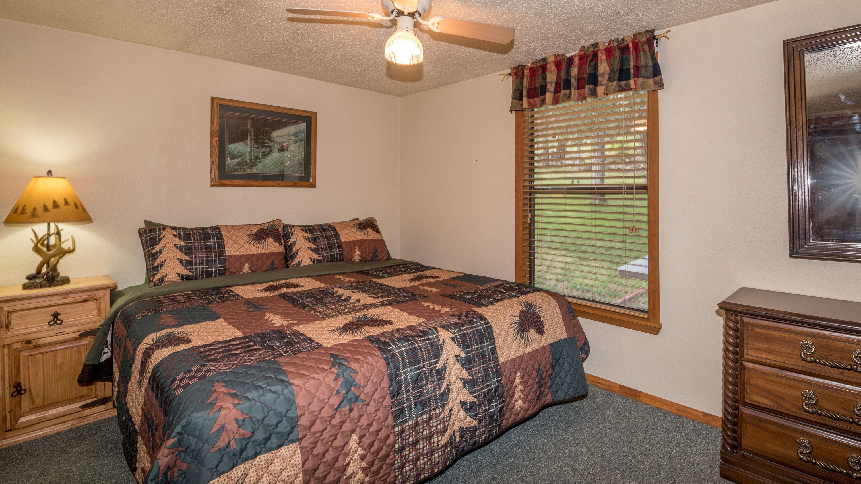 A bedroom with a bed, a nightstand, a dresser, a ceiling fan, a window with blinds, and a framed picture on the wall.