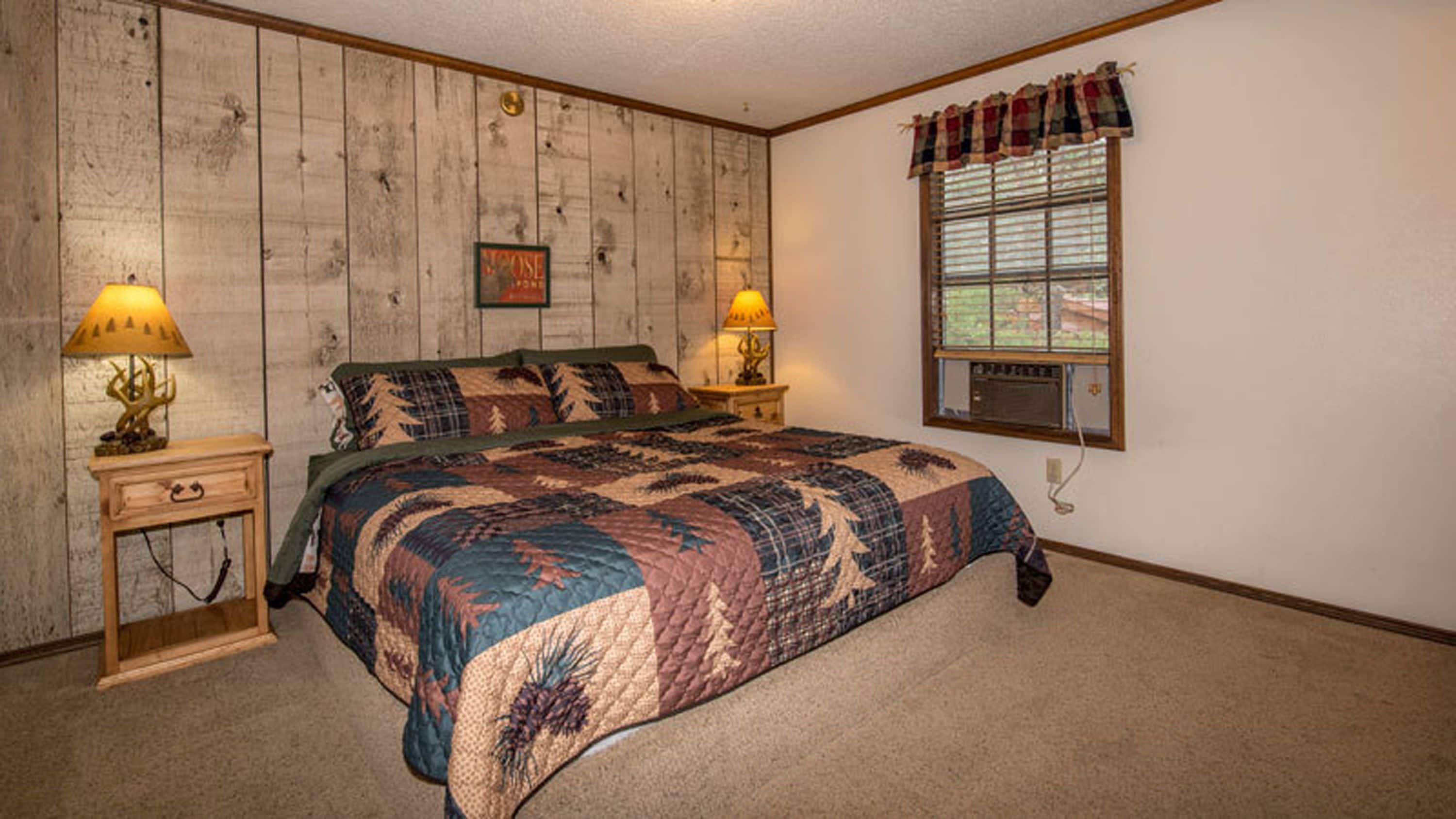 Um quarto com parede revestida de madeira, uma cama grande com colcha, duas luminárias de cabeceira, uma janela com persianas e um ar-condicionado fixado na parede.