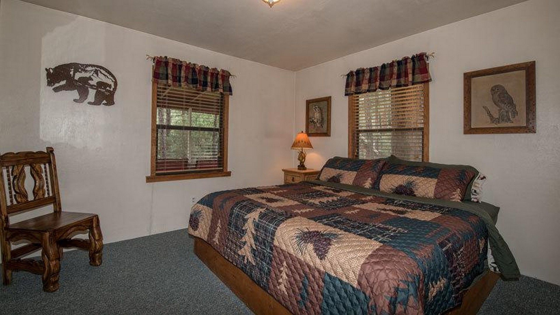 A bedroom with a large bed, two windows with curtains, a chair, and a lamp.