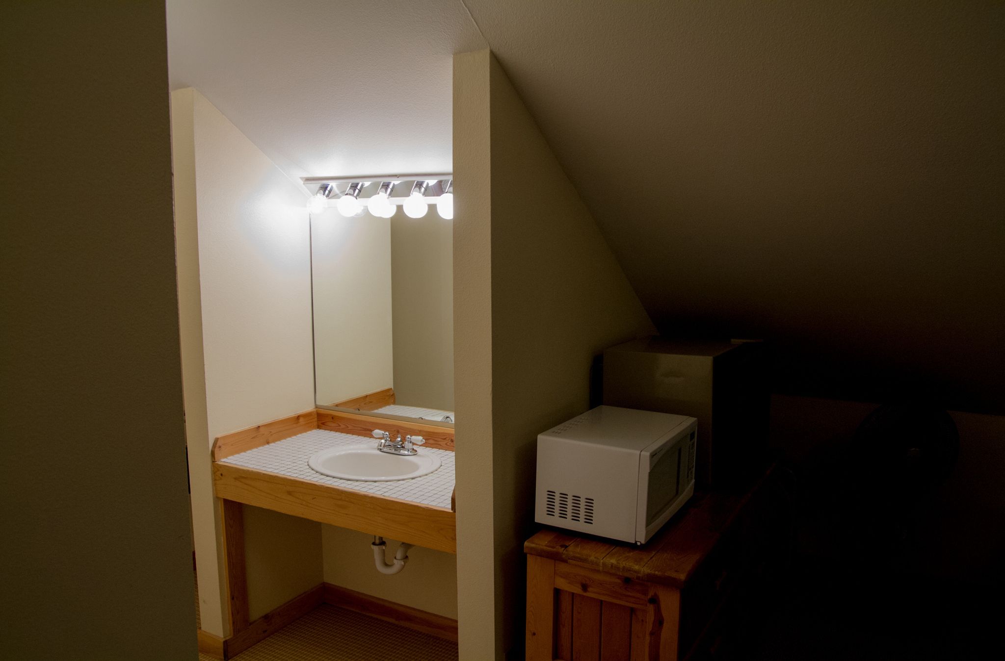 Un baño pequeño y poco iluminado, con un tocador de madera, un lavamanos blanco y un microondas sobre un mueble de madera.