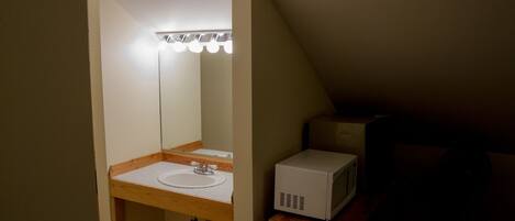 Un baño pequeño y poco iluminado, con un tocador de madera, un lavamanos blanco y un microondas sobre un mueble de madera.