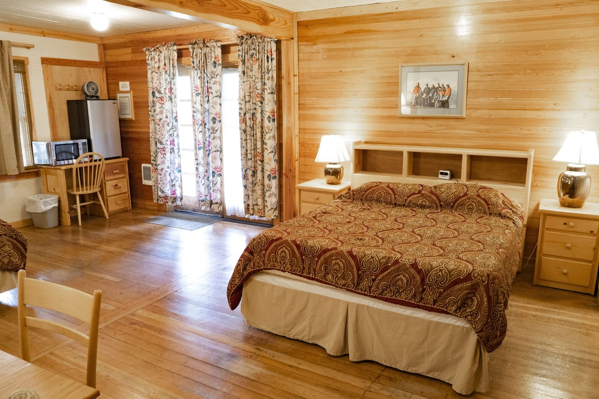 Ein gemütliches Zimmer mit Holzboden, einem Bett mit gemustertem Überwurf, einem Schreibtisch mit Stuhl, einem Fernseher und einem Bild an der Wand.
