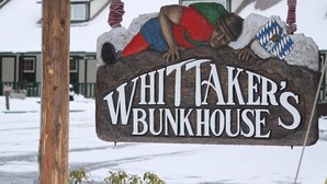 Une pancarte en bois représente un homme vêtu d’une chemise rouge et d’un short bleu, appuy é contre la pancarte. On peut y lire : « WHITTAKER’S BUNKHOUSE ».