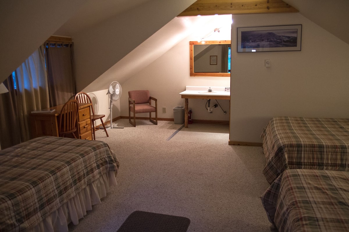 Ein Schlafsaalzimmer mit zwei Betten, einem Schreibtisch, einem Stuhl, einem Ventilator und einem Fenster mit Vorhängen.