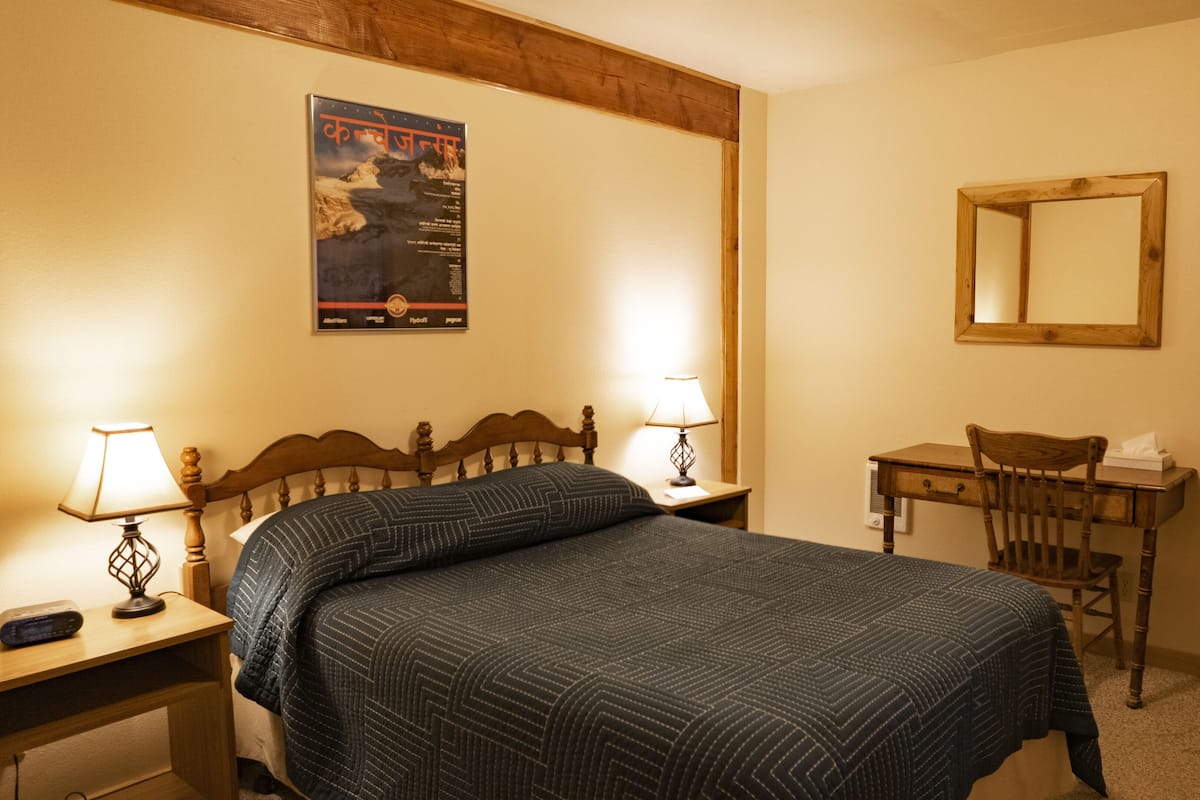 Ein Schlafzimmer mit einem Bett, zwei Nachttischlampen, einem Holznachttisch, einem Schreibtisch, einem Stuhl, einem Spiegel und einem Poster an der Wand.