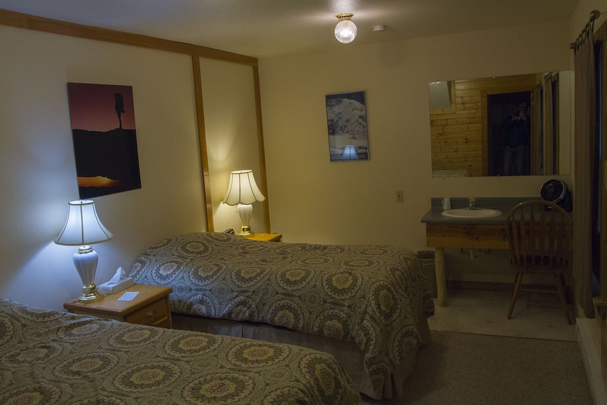 Ein Hotelzimmer mit zwei Betten, einem Nachttisch mit Lampe, einem Spiegel, einem Waschbecken und einem Sessel.
