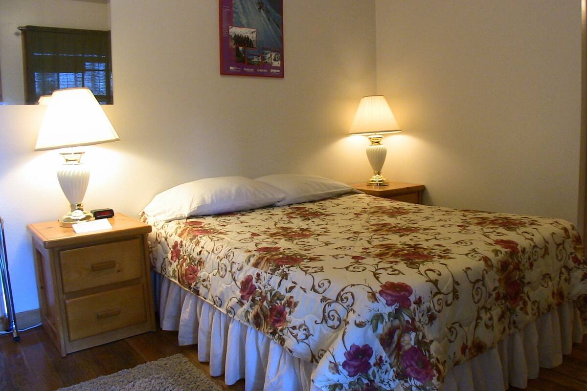 Ein Schlafzimmer mit einem Bett, zwei Nachttischlampen, einem Nachttisch, einer hölzernen Kommode und einem durch eine offene Tür einsehbaren Badezimmer.