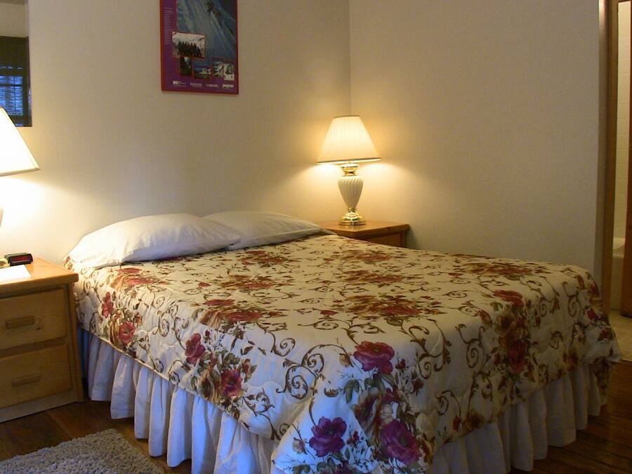 A bedroom with a bed, two bedside lamps, a nightstand, a wooden dresser, and a bathroom visible through an open door.