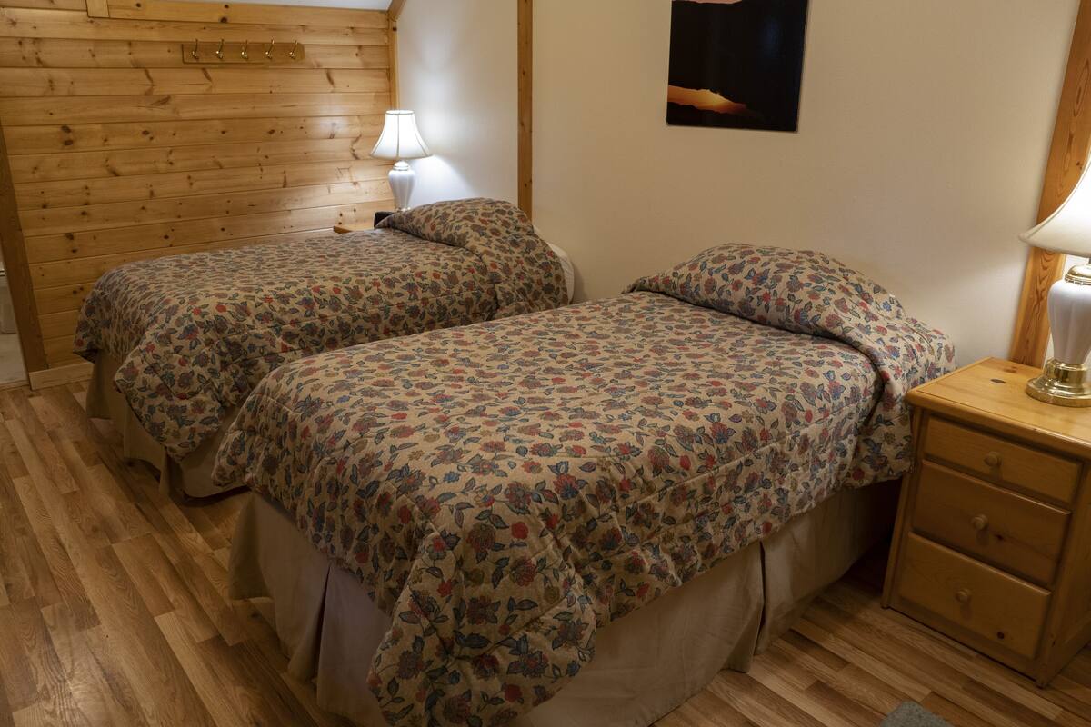 Ein Zimmer mit zwei Betten, Holzboden, einem Nachttisch mit Lampe und einem an der Wand hängenden Bild.
