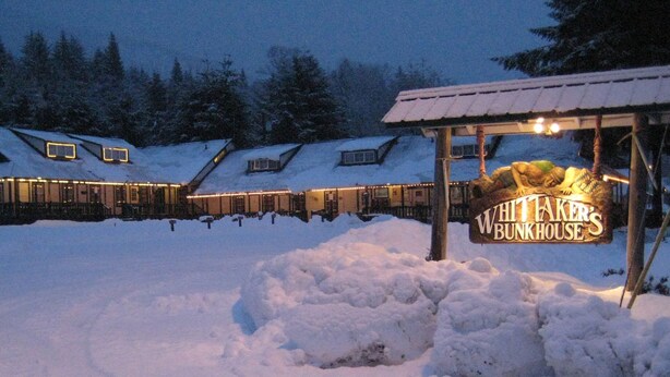 Un bâtiment enneigé, sur lequel on peut lire, sur une enseigne, les mots « Whittaker's Bunkhouse », de nuit.