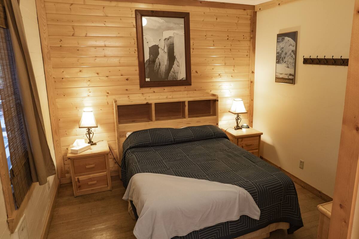 Ein Zimmer mit Holzwänden, einem Bett, zwei Nachttischen mit Lampen und gerahmten Bildern an der Wand.